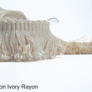 Könnte beinhalten: Nahaufnahme einer cremefarbenen Fransenborte mit dezentem Glanz. Die Borte besteht aus Baumwolle und Rayon und heißt "Cotton Ivory Rayon".
