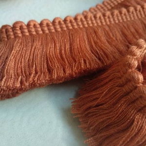 May include: Close-up of a brown tassel trim with a fringe detail. The trim is made of a soft, woven fabric and has a subtle texture.