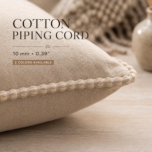 May include: A beige cotton pillow featuring a textured piping cord edge. The text on the image reads: COTTON PIPING CORD, 10 mm • 0.39”, 2 COLORS AVAILABLE. The cord has a repeating beaded appearance along the seam of the fabric.