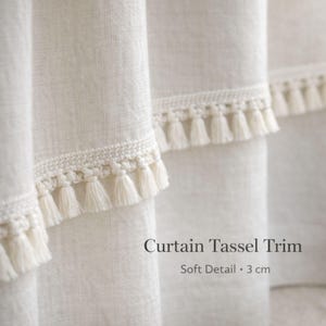 Op de afbeelding: Close-up van off-white gordijnstof met een decoratieve kwastjesbies. De bies heeft een gehaakte rand met kleine, crèmekleurige kwastjes. De tekst "Curtain Tassel Trim" en "Soft Detail • 3 cm" zijn zichtbaar.