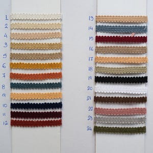 May include: A display of 24 different colored decorative trims, each with a scalloped edge. The trims are arranged in rows on white cards, with colors ranging from cream and beige to navy, burgundy, and olive green. Numbers are written in blue ink next to each trim.
