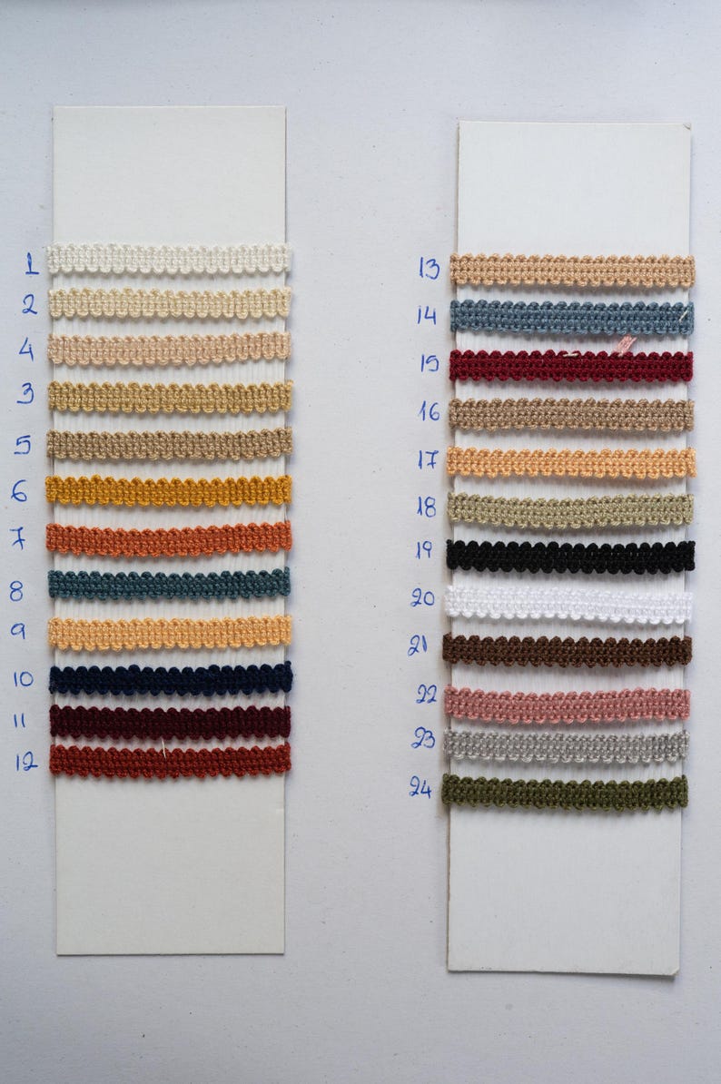 May include: A display of 24 different colored decorative trims, each with a scalloped edge. The trims are arranged in rows on white cards, with colors ranging from cream and beige to navy, burgundy, and olive green. Numbers are written in blue ink next to each trim.