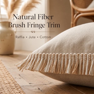 Pode incluir: Uma almofada bege com uma borda de franjas de fibras naturais de ráfia, juta e algodão. O texto na imagem diz: Natural Fiber Brush Fringe Trim, Raffia • Jute • Cotton. A almofada tem uma superfície de tecido com textura visível.