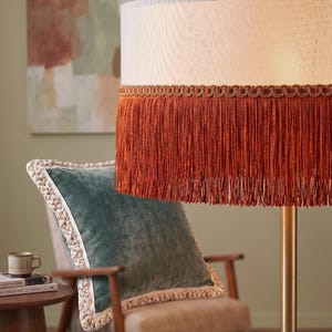 Op de afbeelding: Een vloerlamp met een crèmekleurige lampenkap afgezet met een roestkleurige franje. Een teal fluwelen kussen met een crèmekleurige rand ligt op een houten stoel. Een kleine bijzettafel met een mok en boeken.