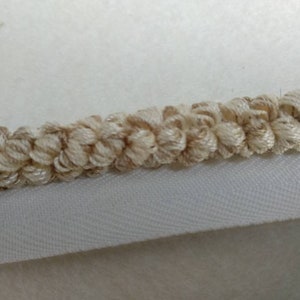 Peut inclure: Gros plan sur une bordure en crochet beige avec une teinte légèrement brun foncé. La bordure est fixée à un fond en tissu blanc.