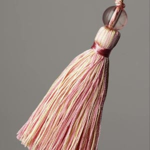 May include: A pink and cream tassel with a clear glass bead at the top. The tassel is made of thin strands of yarn and is tied with a red ribbon.