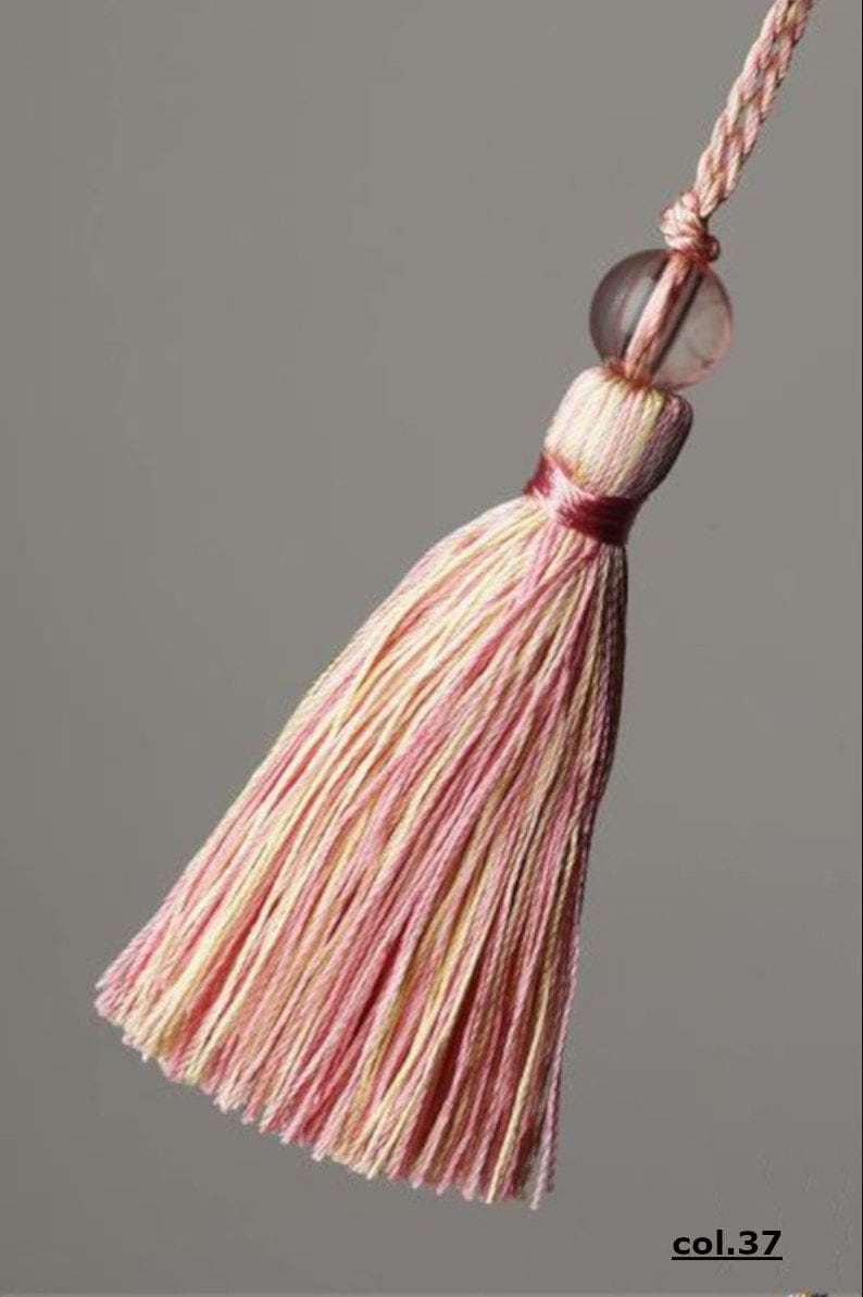 May include: A pink and cream tassel with a clear glass bead at the top. The tassel is made of thin strands of yarn and is tied with a red ribbon.
