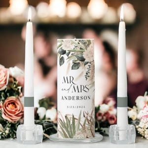 May include: Two white pillar candles with gray bands, one with a floral design and the text "MR and MRS ANDERSON 5/23/2024" on the side, on a clear glass base.