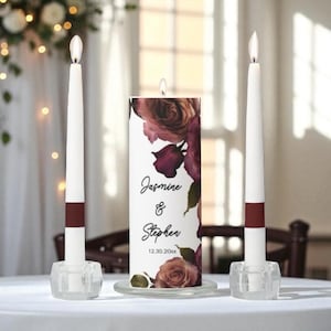 May include: A wedding candle set featuring a central pillar candle with floral design and the names "Jasmine & Stephen" and date. Two white taper candles with burgundy bands and clear holders flank the pillar candle.