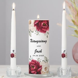 May include: Two white unity candles with burgundy and cream floral designs. The larger candle has the text "Xiangxiang and Josh 09.24.2022" printed on it.