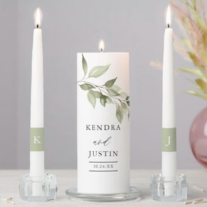 May include: A white unity candle set with two taper candles. The center candle has a watercolor leaf design and the names "Kendra and Justin" with a date. The taper candles have initial stickers.