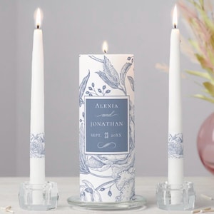 May include: A set of three lit candles. Two white taper candles with blue floral bands flank a larger pillar candle. The pillar candle features the names "Alexia and Jonathan" and the date "Sept. 23 20XX" in a blue frame, surrounded by a blue floral design.