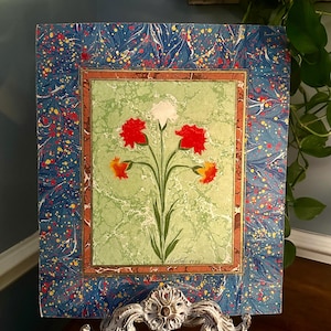 May include: A framed floral artwork featuring a central image of red, white, and yellow flowers with green stems against a light green background. The artwork is framed with a blue marbled border with red and yellow speckles. The artwork is displayed on a decorative white stand.