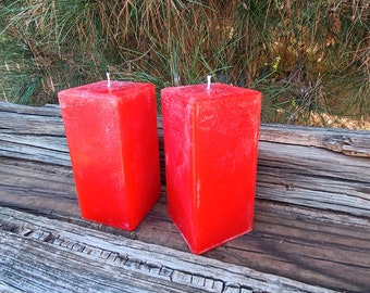 May include: Two vibrant red square pillar candles with white wicks. The candles have a textured, slightly frosted appearance. They are displayed on a weathered wooden surface, with green foliage in the background. These decorative candles are suitable for home decor.