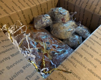 May include: A brown cardboard box filled with white packing paper and gold ribbon. The box contains a loaf of bread and several blueberry muffins wrapped in plastic.