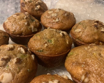 May include: A close-up shot of a dozen golden-brown muffins, each topped with chopped walnuts. The muffins appear moist and have visible flecks of green, suggesting zucchini. They are arranged in a clear container.