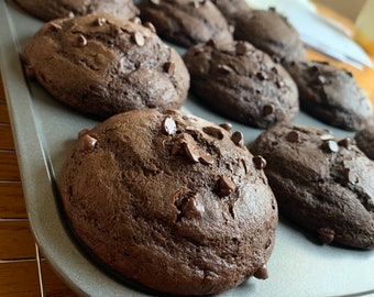 May include: A baking sheet filled with freshly baked chocolate chip muffins. The muffins are a deep brown color and have a slightly domed top. The muffins are arranged in a single layer on the baking sheet.