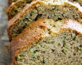 Puede incluir: Primer plano de una hogaza de pan de calabacín cortado en rebanadas gruesas. El pan es de color marrón claro y tiene una textura ligeramente desmenuzable. El calabacín es visible en todo el pan, lo que le da un aspecto moteado verde.