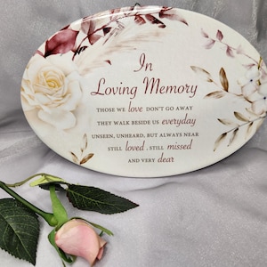May include: Oval memorial plaque with floral design and the inscription "In Loving Memory." The design features a cream background, burgundy and cream floral elements, and a light pink rose. A single rose is in the foreground.