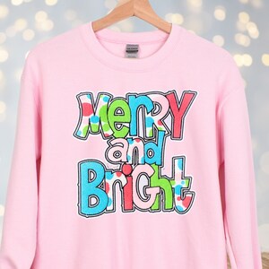 May include: A light pink sweatshirt with the words "Merry and Bright" in a colorful, playful font. The text has a tie-dye effect with green, blue, and red accents. The sweatshirt is displayed on a wooden hanger.