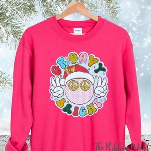 May include: Pink Christmas sweatshirt with a groovy peace sign wearing a Santa hat and the text "Groovy & Bright".