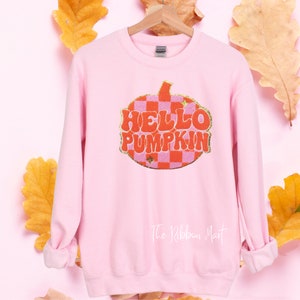 May include: Pink sweatshirt with a checkered pumpkin graphic and the text "HELLO PUMPKIN" in a retro font. The sweatshirt has a crew neck and long sleeves. The text "The Ribbon Mart" is printed on the bottom of the sweatshirt.
