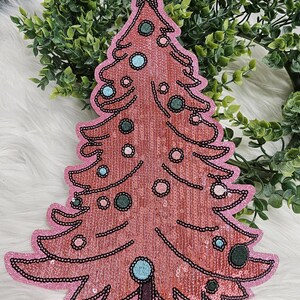 May include: A pink sequined Christmas tree decoration with a star on top. The tree is outlined in black and decorated with blue and green circular ornaments. The tree is on a white, fluffy surface.