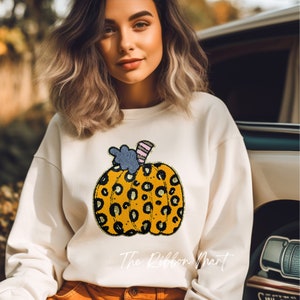 May include: A white sweatshirt with a yellow and black leopard print pumpkin design. The pumpkin has a blue and pink stem. The text "The Ribbon Mart" is printed on the bottom of the sweatshirt.