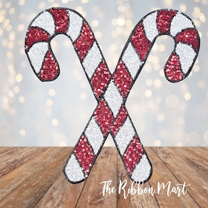 May include: Two red and white candy cane ornaments with a sparkly finish. The candy canes are crossed over each other and are on a wooden surface. The text "The Ribbon Mart" is visible at the bottom of the image.