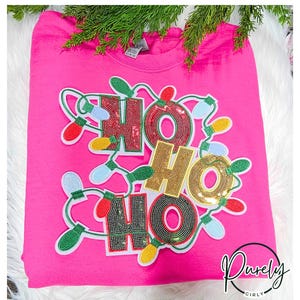 May include: Pink t-shirt with a sparkly "Ho Ho Ho" design featuring Christmas lights. The design is made of sequins and is surrounded by a string of colorful lights.