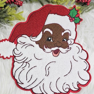 May include: A festive Christmas decoration featuring a smiling Santa Claus face. The design includes a red hat and outline, a white beard, and a brown face. A sprig of holly with red berries adds a touch of holiday cheer.