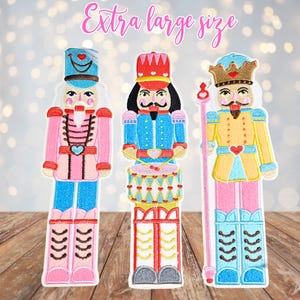 May include: Three extra large embroidered nutcracker figures in pink, blue, and yellow. Each figure has a unique design, including hats, uniforms, and accessories. The text "Extra large size" is above the figures.