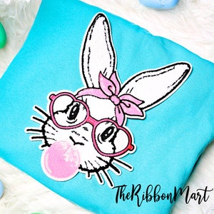May include: A white bunny with pink glasses and a pink bow is blowing a pink bubble. The bunny is on a turquoise blue background.