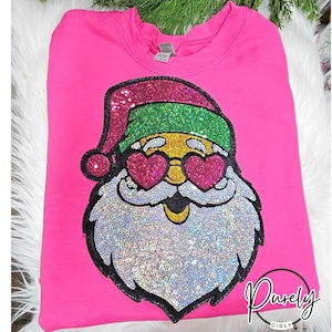 May include: A bright pink sweatshirt featuring a sequined Santa Claus design. The Santa has heart-shaped sunglasses, a glittery hat, and a sequined beard. The sweatshirt has the text "Purely Girly" in a cursive font.