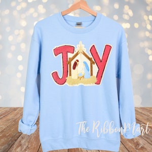 May include: A light blue sweatshirt with the word "JOY" in red letters. The letter "O" is replaced with a nativity scene. The sweatshirt is hanging on a wooden hanger against a blurred background.