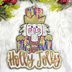 May include: A sequined Christmas decoration in the shape of a nutcracker soldier. The nutcracker has a gold crown and a red and gold uniform. The words "Holly Jolly" are written in sequins below the nutcracker.