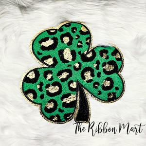 Puede incluir: Trébol verde con un diseño de estampado de leopardo dorado y negro. El trébol está hecho de fieltro y tiene un acabado brillante.