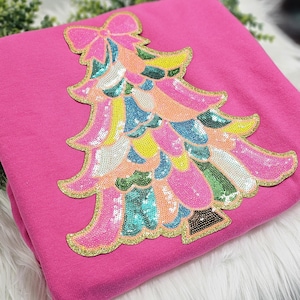 May include: A bright pink sweatshirt featuring a colorful sequined Christmas tree design. The tree is adorned with sequins in shades of pink, blue, yellow, and green, outlined with gold glitter. The bow at the top adds a festive touch.