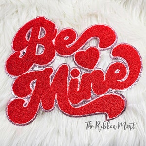May include: Red and pink glitter iron-on patch with the words "Be Mine" in a retro font. The patch has a heart shape in the middle of the word "Mine".