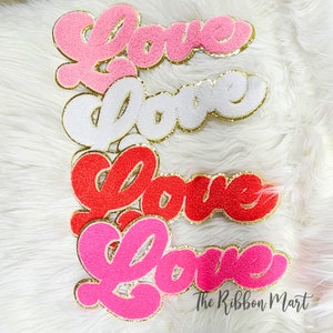 May include: Four fuzzy "Love" patches in pink, white, red, and hot pink with gold glitter trim. The patches are arranged in a stack, with the pink patch on top and the hot pink patch on the bottom. The patches are on a white fluffy surface. The text "The Ribbon Mart" is visible in the bottom right corner of the image.