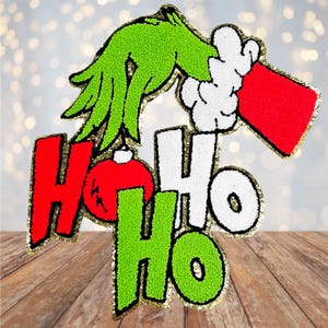 May include: Green, white, and red felt Christmas ornament with the words "Ho Ho Ho" and a green hand holding a red ornament.