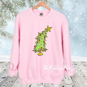 May include: Pink sweatshirt with a green Christmas tree graphic with colorful ornaments and a gold star on top. The text "The Ribbon Mart" is visible at the bottom of the image.