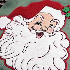 May include: Embroidered Santa Claus face with a red hat and beard, outlined in red. The Santa Claus face has rosy cheeks and a smiling expression. A sprig of holly is visible near the hat. The design is on a green background.