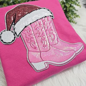 May include: A pink sweatshirt featuring a sequined Santa hat and a pink sequined cowboy boot design. The Santa hat is red with white trim and a white pom-pom. The cowboy boot has intricate sequin detailing.