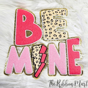 May include: Pink and red felt letters spelling "BE MINE" with a gold glitter outline. The "E" is a leopard print design and the "M" has a pink lightning bolt with a leopard print design.