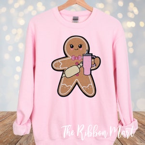 May include: A light pink sweatshirt featuring a gingerbread man graphic. The gingerbread man is holding a rolling pin and a pink tumbler. The design has a black outline and a pink bow tie. The text "The Ribbon Mart" is at the bottom.