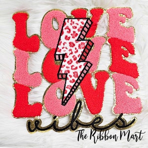 May include: A decorative patch with the words "LOVE VIBES" in red and pink, with gold trim. A lightning bolt with a leopard print design is in the center. The Ribbon Mart logo is in the bottom right corner.