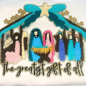 May include: Cream-colored decorative pillow with an embroidered Nativity scene. The design features figures in blue, pink, and teal, with gold accents and the text "the greatest gift of all".