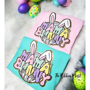 May include: Two pastel colored sweatshirts with a glittery, pastel colored "Mama Bunny" design with bunny ears. The design is in a circle with gold glitter around the edge. The sweatshirts are on a white fluffy surface with Easter eggs.