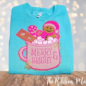May include: A turquoise sweatshirt features a pink and gold embroidered design of a gingerbread person in a mug of marshmallows. The mug reads "MERRY & BRIGHT" in pink lettering. A candy cane and chocolate bar are also in the mug.